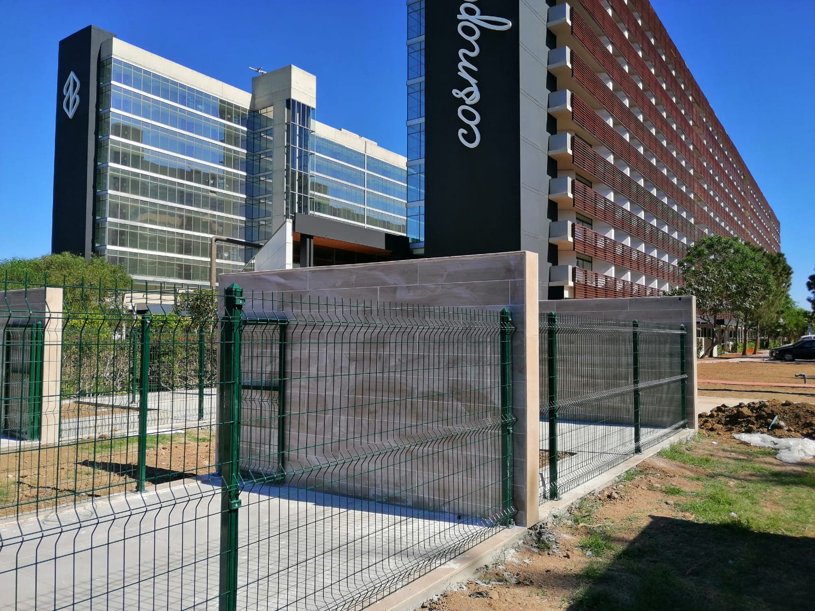 Green welded mesh fence mounted on a low wall in front of the Cosmopolitan hotel building